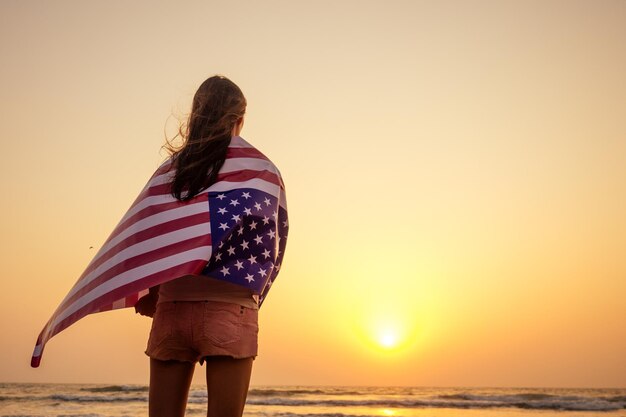 View from the back young brunette female person holding national American flag cover ocean beach holiday travel at summer romantic sunset .America independence day concept.