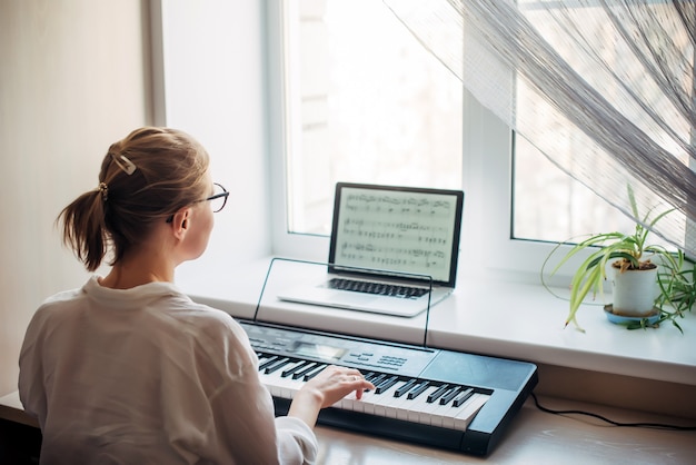 Вид сзади молодая женщина играет на синтезаторе, читает заметки на экране ноутбука. Самостоятельное обучение игре на фортепиано в домашних условиях. Увлечение музыкой, хобби, отдых, саморазвитие.