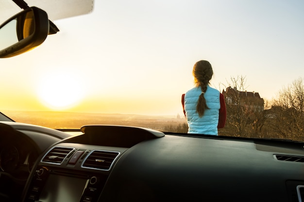 暖かい夕日の景色を楽しんでいる彼女の車の近くに立っている若い女性の中からの眺め。夜の地平線を見ている車のボンネットに寄りかかって女の子の旅行者。