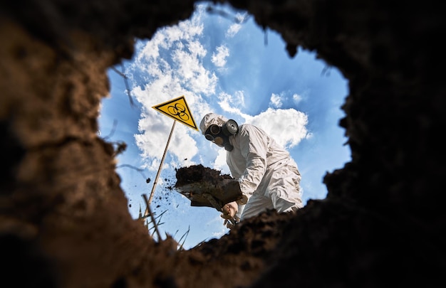 View from inside pit male ecologist digging pit by shovel wearing protective overalls and gas respirator near biohazard symbol warning about dangerous biological materials