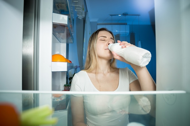 View from inside of refrigerator on young woman drinking milk at night