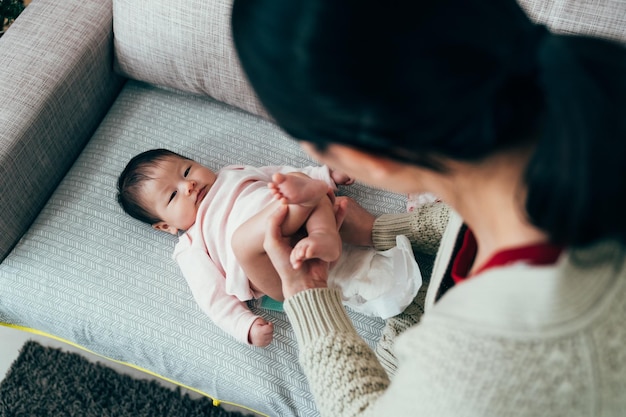 Vista dall'alto adorabile bambino sta fissando la custode femminile. la mamma asiatica per la prima volta sta alzando i piedi della figlia e le cambia il pannolino. stile di vita autentico