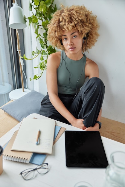 View from overhead of curly haired young woman poses in modern\
quiet workspace uses tablet connected to earphones listens audio\
course learns foreign language online looks seriously at\
camera