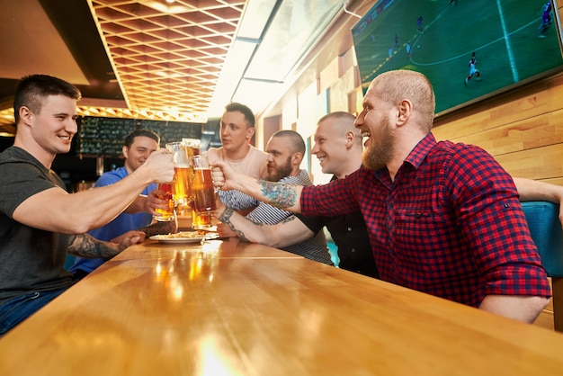 Foto vista dal lato della compagnia maschile allegra che riposano insieme nel pub nei fine settimana. uomini felici che bevono birra, tostano, ridono e parlano al bar. concetto di felicità e divertimento.