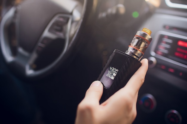 View from the side of a young man smoking an e-cigarette as he drives his car on an urban street Holds and adjusts the power of the electronic cigarette