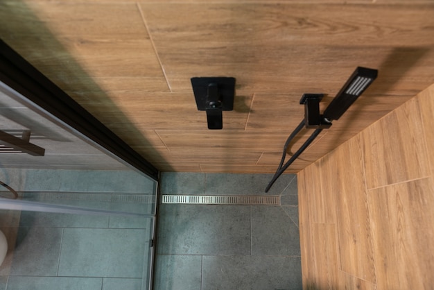 Вид сверху кабины на душ с деревянными плиточными стенами и серым кафельным полом со сливом из металла мимо смесителя и насадки для душа