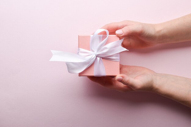 View from the top a woman's hand holds a pink gift box with a white bow on a pink background. A place to copy.