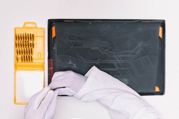 View from above of a white gloved hands of an engineer unscrewing the screws to open the lid of a laptop before being repaired on a white table