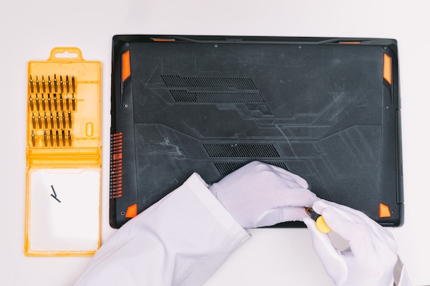 View from above of a white gloved hands of an engineer unscrewing the screws to open the lid of a laptop before being repaired on a white table