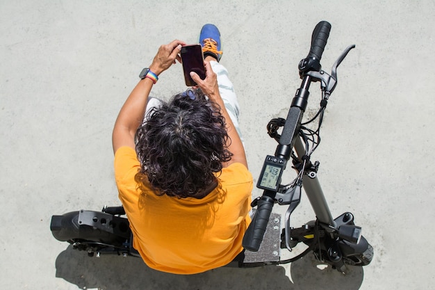 Foto vista dall'alto di una donna seduta sull'asfalto usando il suo smartphone accanto a uno scooter elettrico