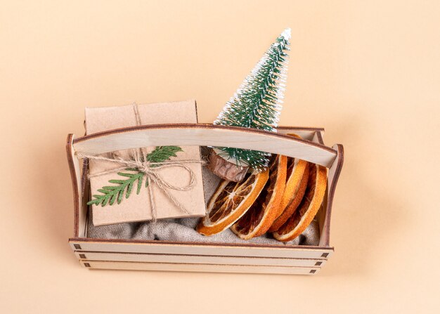 Vista dall'alto alla scatola di legno con albero di natale decorativo, fette d'arancia secche e confezione regalo su beige.