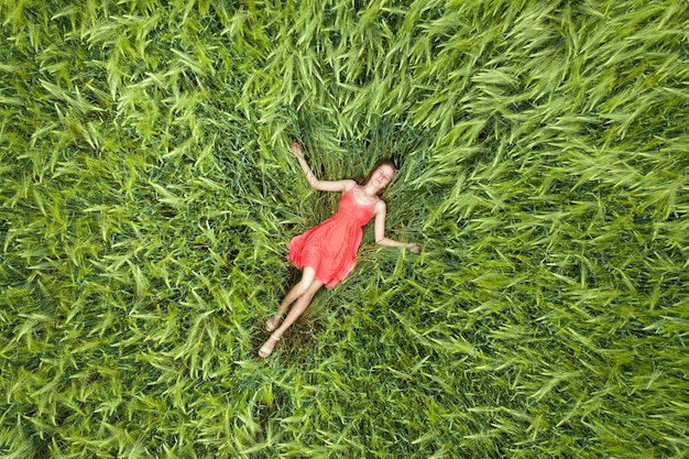 View from above of young attractive slim woman in red dress laying with closed eyes in green wheat field.