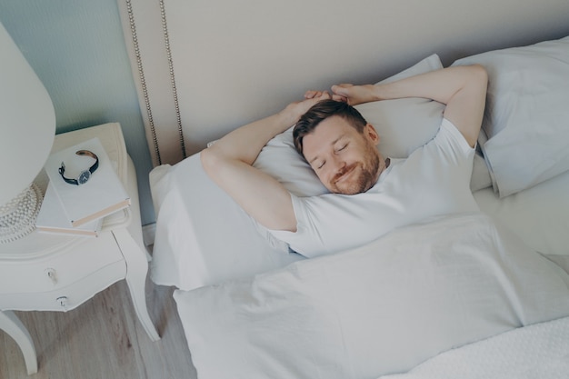 白い寝具で居心地の良いベッドに横たわって、魅力的な笑顔で眠っている若い幸せなリラックスした白人男性の上からの眺め。健康管理おやすみ睡眠と休息の概念