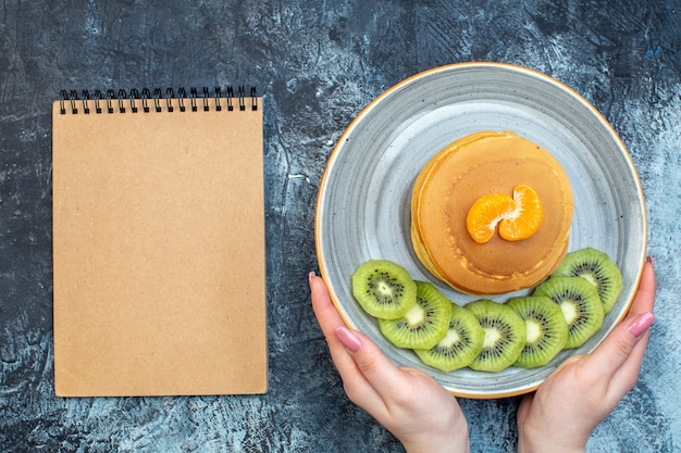 Above view of hand holding fluffy American-style pancakes made with natural yogurt served with kiwifruit and tangerine on a plate and notebook on ice background with free space