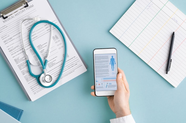 Foto sopra la vista delle mani del medico con i gadget che scorrono le statistiche mediche tra i documenti negli appunti con lo stetoscopio e la carta vuota