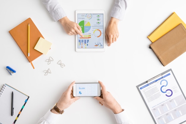 Photo view of hands of two contemporary analysts or brokers analyzing financial data while using touchscreen and smartphone in front of each other
