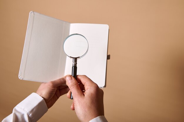 Una vista di un'immagine del foglio bianco bianco con una lente d'ingrandimento o una lente d'ingrandimento in mani femminili sulla parete beige con lo spazio della copia