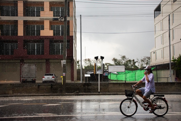 タイのノンタブリーのバンブアトン市で雨滴が嵐を降らせている間、タイの人々が路上で乗馬や自転車を運転している風景の田園地帯と交通道路を表示します