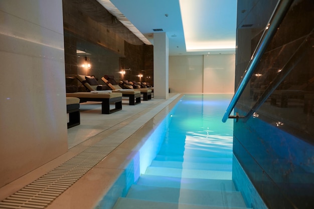 View of the luxurious modern interior of the spa and wellness center with sun loungers by the thermal pool with underwater lighting and a waterfall.