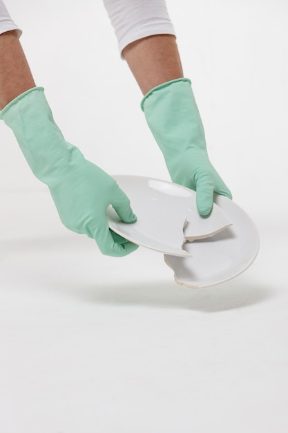 View of man hands in protective rubber gloves picking up a broken plate from the floor during cleaning or after a quarrel. Isolated on white background. Cleanup concept.