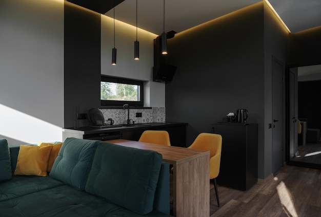 View of modern studio apartment with green sofa and yellow dining chairs in dark kitchen