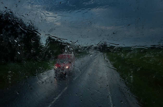 Вид встречной машины на трассе с включенными фарами через стекло, мокрое от проливного дождя в каплях и каплях в очень плохую погоду вечером