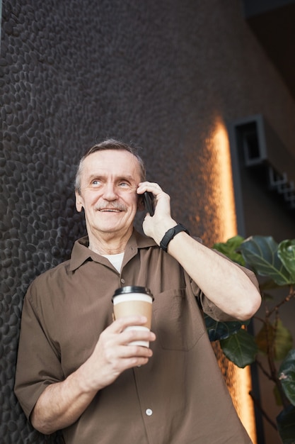 Photo below view of positive confident senior caucasian man in brown shirt leaning on black stone-textured wall and communicating by cellphone