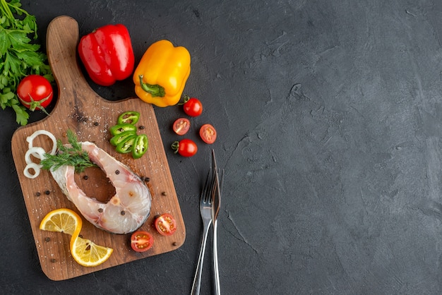 Above view of raw fish and fresh chopped vegetables lemon slices spices on a wooden board cutlery set green bundle on the right side on black distressed surface