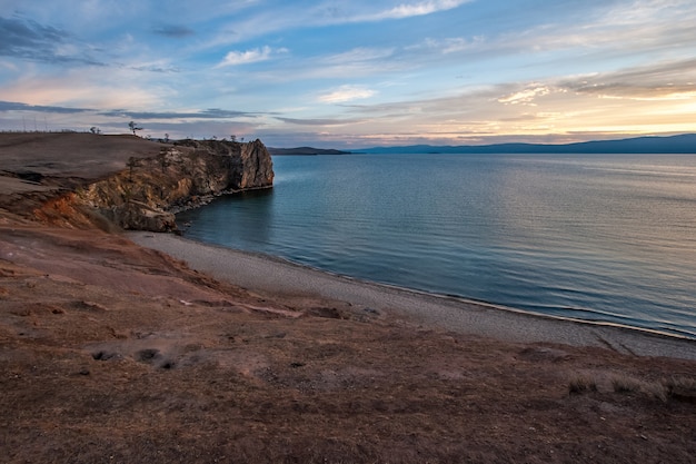 Vista del promontorio roccioso e del lago baikal sull'isola di olkhon al tramonto. acqua blu e cielo con nuvole. riva sabbiosa. colori pastello.