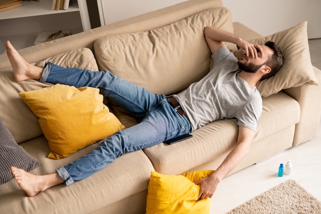 Sopra la vista del giovane uomo barbuto triste perplesso in abbigliamento casual sdraiato sul divano nel soggiorno e che copre il viso con la mano in quarantena