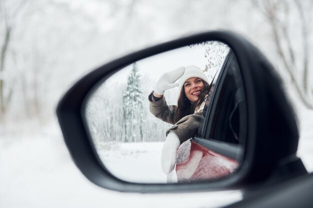 Photo view in the side mirror beautiful young woman is outdoors near her red automobile at winter time