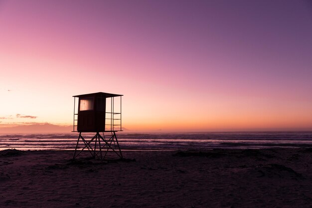 Photo view of silhouette lifeguard hut at sandy beach against scenic view of sea and clear purple sky