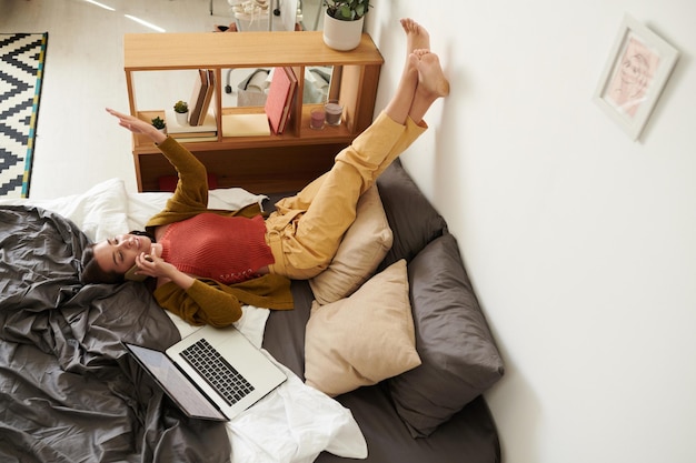 Photo above view of smiling young woman in yellow pants lying on bed with laptop and chatting by mobile phone