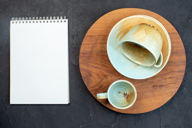 Above view of soft colorful cups and saucer on brown round cutting board next to notebook on black background with free space