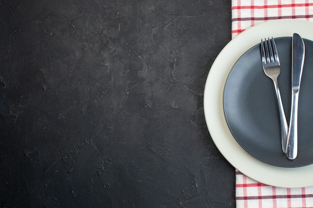 Above view of stainless cutlery set on dark gray color and white empty plates on red stripped towel on the left side on black background with free space