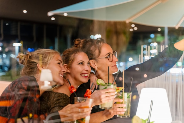 Вид через стакан женщин, делающих селфи и пьющих коктейли в ресторане ночью