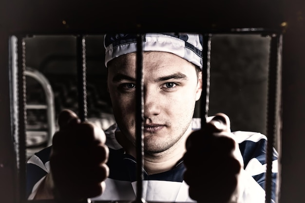 View through iron door with prison bars on young prisoner holding bars wearing prison uniform in a jail cell