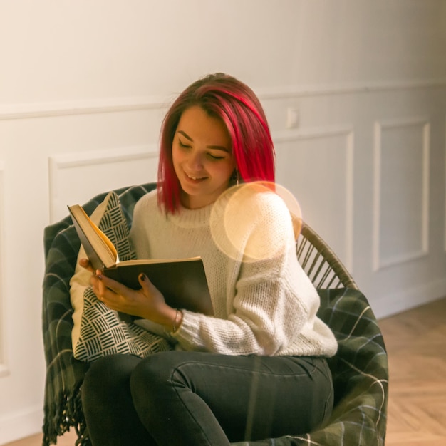 Photo view through a window with garlands of a dreamy charming young lady in a sweater reading a book in a rocking chair