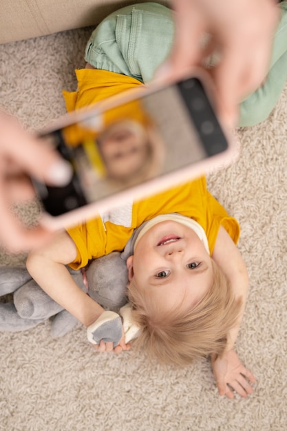床におもちゃで横たわっている幸せなかわいい息子の写真を撮っている間スマートフォンを使用して認識できない親のビューの上