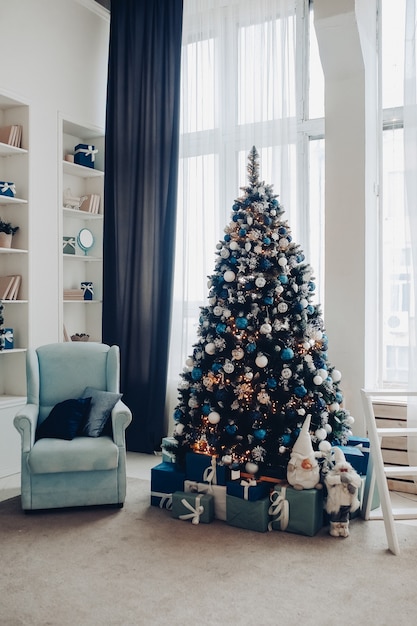 View over white modern room decorated for Christmas holidays. Decorated and illuminated Christmas tree, fireplace with fir branch, candles and handmade paper stars in fireplace.