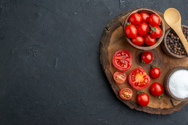 Above view of whole cut fresh tomatoes and spices on wooden board on the left side on black surface with free space