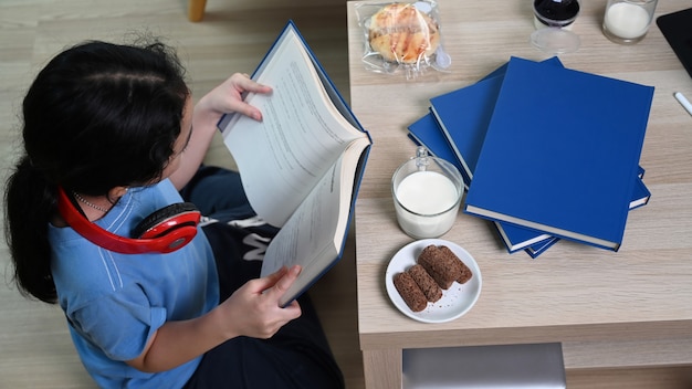 Above view of young asian girl reading book while sitting on floor in living room