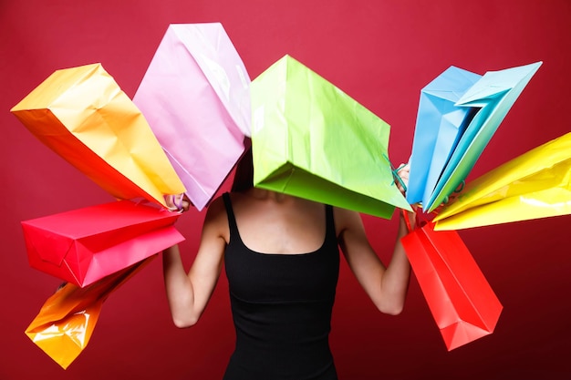 View of young attractive woman with bright lipstick in black dress shaking colorful paper shopping bags on red background she is not visible behind the bags black friday concept