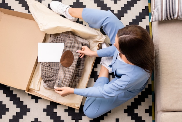 Вид молодой женщины в синей пижаме, сидящей на полу в гостиной, распаковывающей и разворачивающей клетчатую куртку, заказанную в интернет-магазине