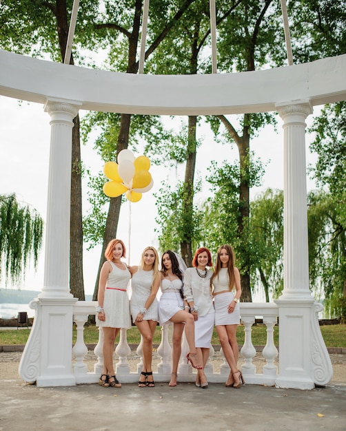 Vijf vrouwen met ballonnen bij de hand droegen op witte jurken op vrijgezellenfeest tegen witte zuilen van boog.