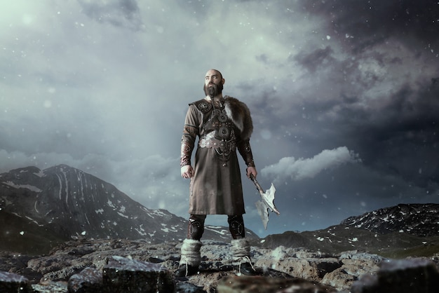 Viking with axe dressed in traditional nordic clothes standing in rocky mountains. Scandinavian ancient warrior