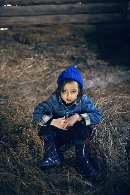 Фото Деревенский мальчик в сапогах сидит на сене в амбаре в теплой одежде и синей шляпе