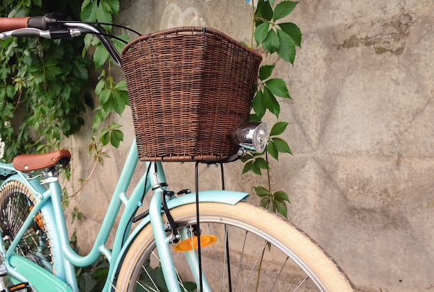 Vintage bicycle with wicker basket and headlamp stands near a concrete fence with green plants.