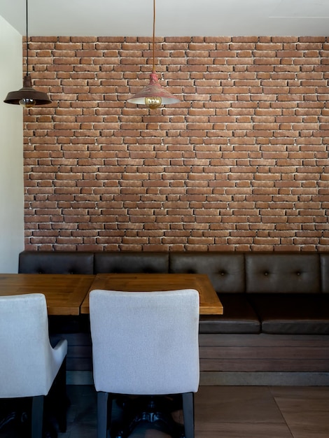 Винтажный потолочный светильник с лампочкой, висящей над пустым современным креслом из серой ткани, длинным кожаным диваном в стиле ретро и деревянным столом, обеденным столом на фоне кирпичной стены в вертикальном стиле