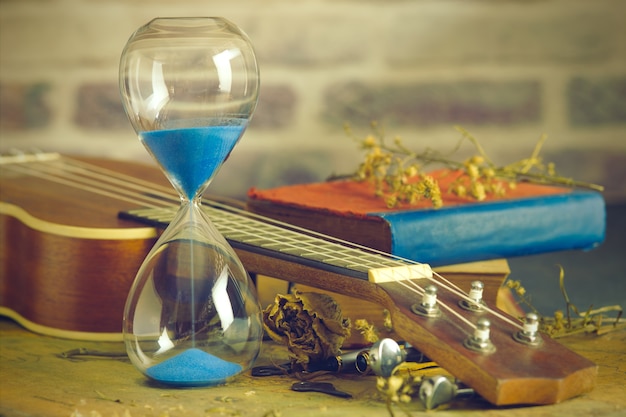 A vintage hourglass and ukulele with an old book and brass pen on a wooden table